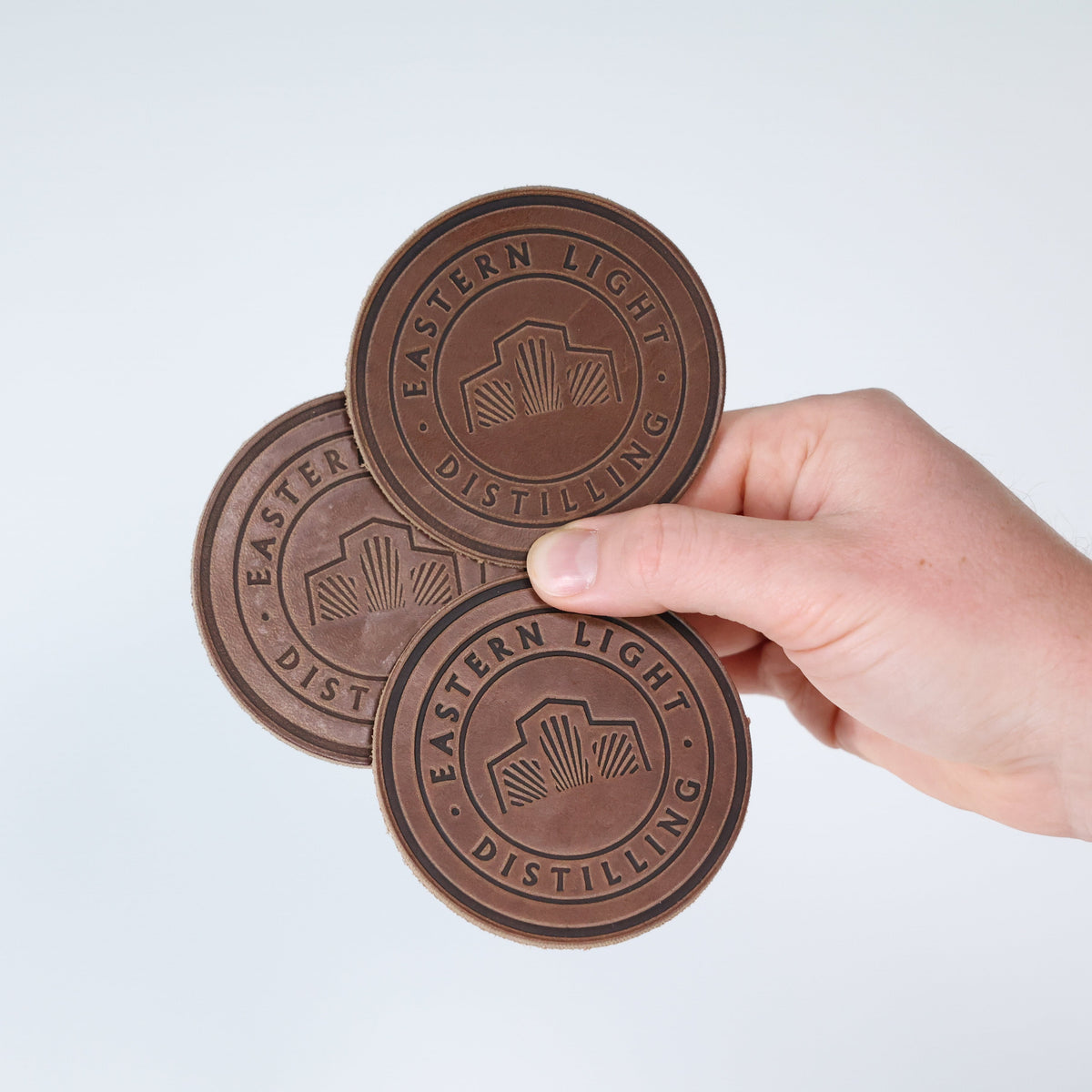 Clayton and Crume Leather Coaster Set of Four for Eastern Light Distilling - Hand holding 3 Coasters on White Backdrop