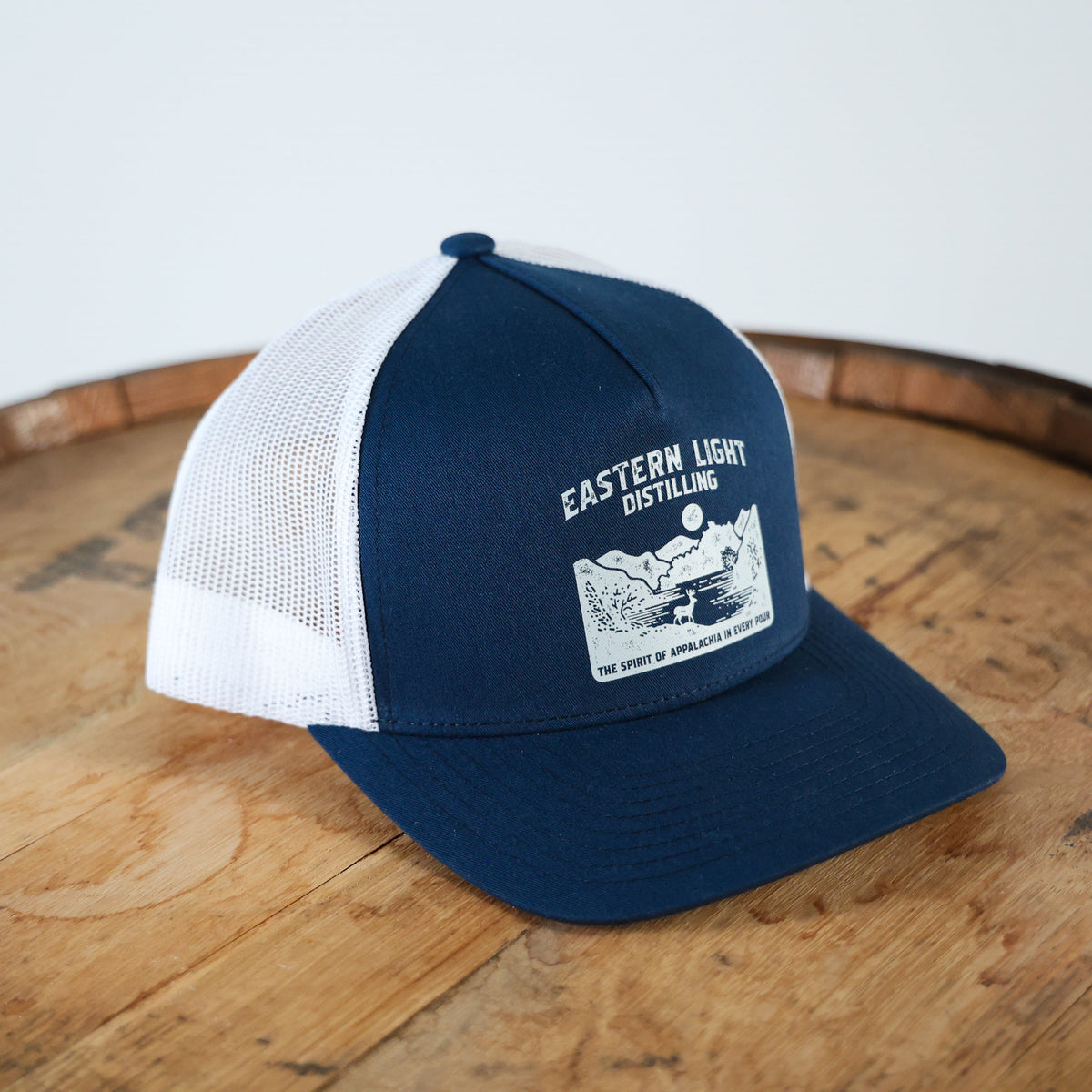 Navy Blue and White Trucker Hat with Eastern Light Distilling Graphic printed on front sitting on Bourbon Barrel